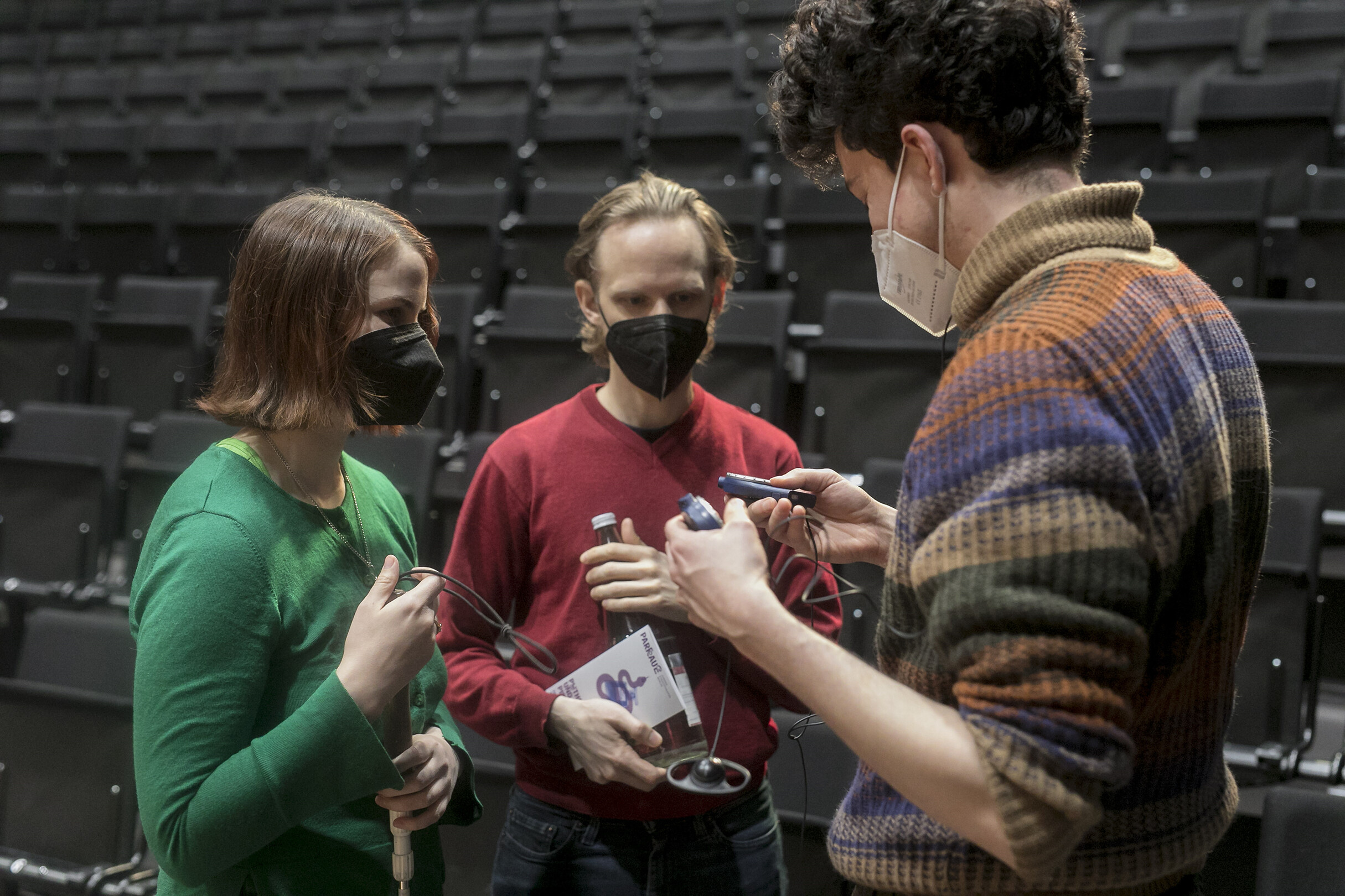 Zwei Personen stehen einer dritten gegenüber, sie tragen alle eine Maske. Hinter ihnen ist ein Zuschauerbereich mit vielen schwarzen Sitzen. Links steht eine Person mit braunem, schulterlangem Haar und grünen Oberteil, die einen Blindenstock in der Hand hält. Rechts von ihr steht eine Person mit blondem, kurzem Haar und einem roten Oberteil. In der einen Hand hält sie einen Flyer, in der anderen eine Glasflasche. Die beiden schauen auf die kleinen Geräte, die die Person zur rechten in der Hand hält. Diese trägt ein mehrfarbiges, gestreiftes Oberteil mit Rollkragen.